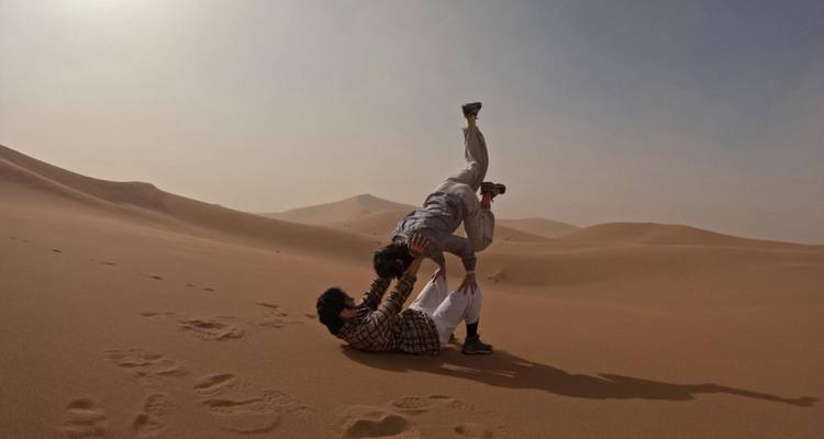 Twee reizigers voeren acro-yoga uit op een uitgestrekte zanduin met wazig woestijngebergte op de achtergrond.