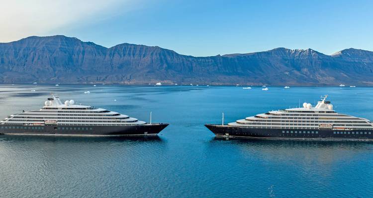 Two sleek expedition yachts face each other on calm blue Arctic waters with mountains behind.
