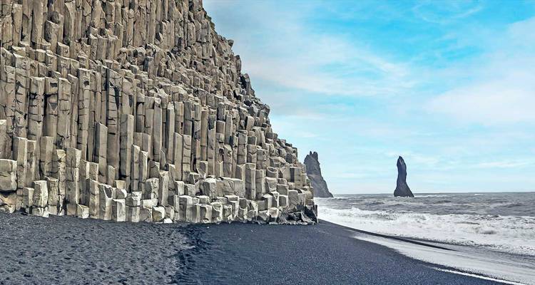 Basaltsäulen-Klippen und schwarzer Sandstrand bei Reynisfjara mit Felsnadeln vor der Küste unter blauem Himmel.
