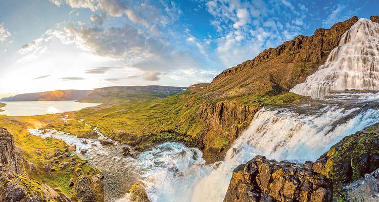 Panoramic view of Dynjandi waterfall cascading down rocky cliffs into green valley at sunset.