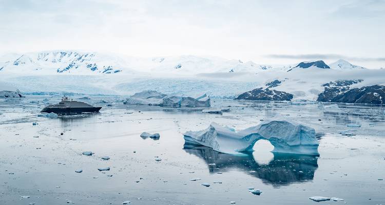 A luxury expedition ship navigates icy Antarctic waters filled with icebergs against snow-covered mountains.