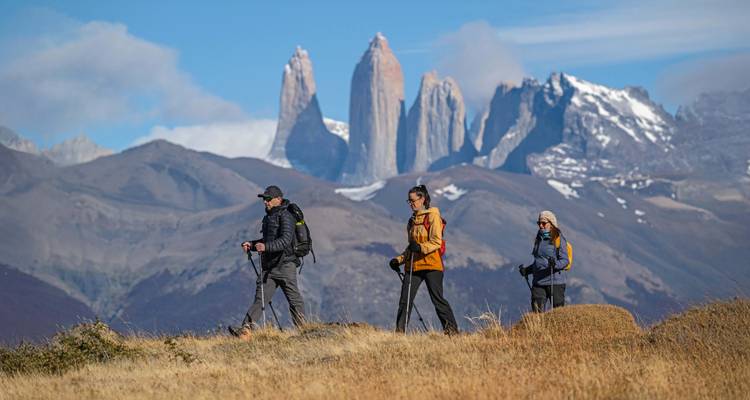 Drei Wanderer mit Stöcken wandern über eine Grasebene zu den dramatischen Granittürmen von Torres del Paine.