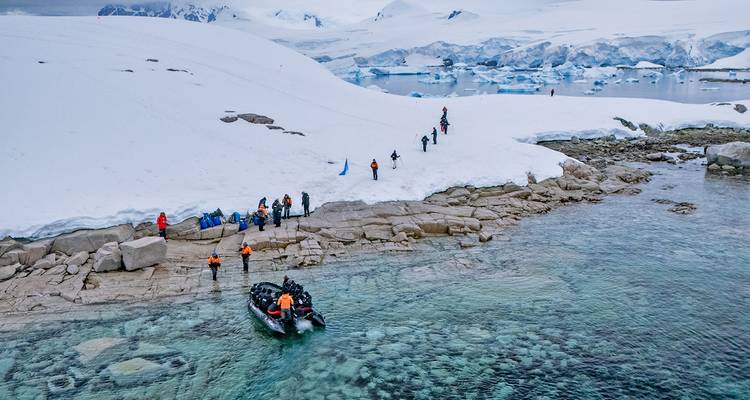 Les passagers de l'expédition en parkas orange débarquent des zodiacs sur un rivage antarctique couvert de neige à côté d'une eau turquoise cristalline.