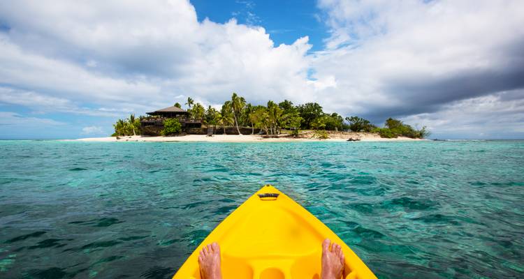 Vue depuis l'avant d'un kayak jaune avec les pieds du pagayeur pointés vers une minuscule île bordée de palmiers dans un lagon turquoise cristallin.