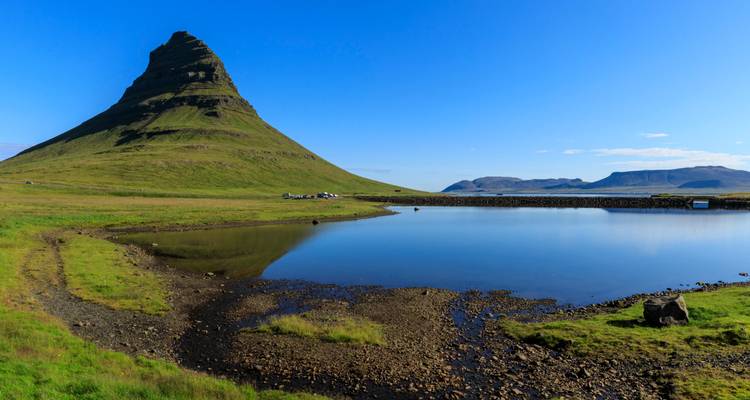 Der ikonische kegelförmige Berg Kirkjufell spiegelt sich in einem stillen Teich unter einem klaren blauen isländischen Himmel wider.