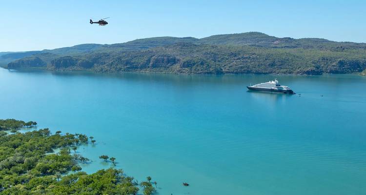 Bahía turquesa rodeada de colinas verdes con un yate de expedición anclado y un helicóptero volando por encima.