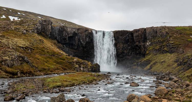 Une puissante cascade se précipite sur une falaise rocheuse dans un ruisseau brumeux entouré de toundra verte et brune sous un ciel islandais couvert.