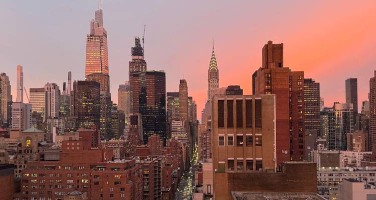 New Yorker Skyline in der Abenddämmerung mit rosa-orangenem Himmel und beleuchteten Wolkenkratzern einschließlich des Chrysler Building.