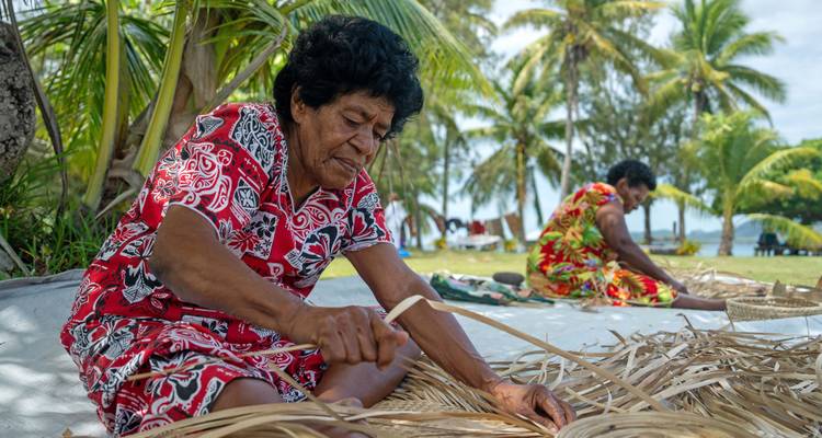 Des femmes en robes colorées tissant des frondes de palmier sous des cocotiers près d'une plage tropicale
