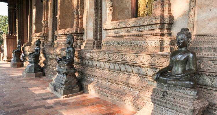 Reihe sitzender Bronze-Buddha-Statuen entlang eines kunstvoll geschnitzten Tempelkorridors mit warmem Licht.