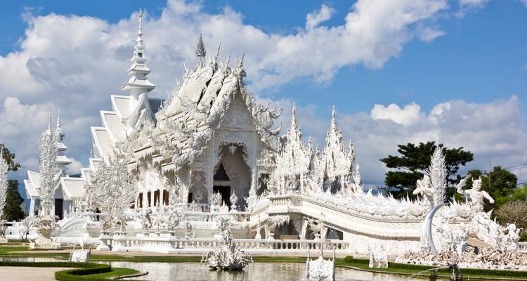 Blendend weißer Wat Rong Khun Tempel mit kunstvollen Skulpturen, der sich in einem Teich unter strahlend hellem Himmel spiegelt.
