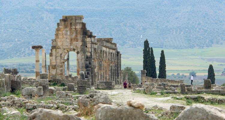Steinbogen und verstreute Ruinen von Volubilis, eingerahmt von Zypressen und Ackerland.