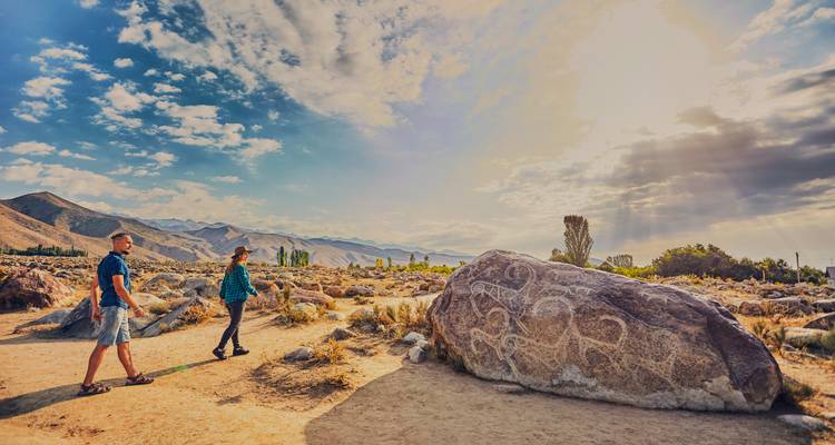 Zwei Reisende wandeln zwischen alten Petroglyphen-Felsen mit dramatischen Bergen und sonnenbeschienenen Wolken.