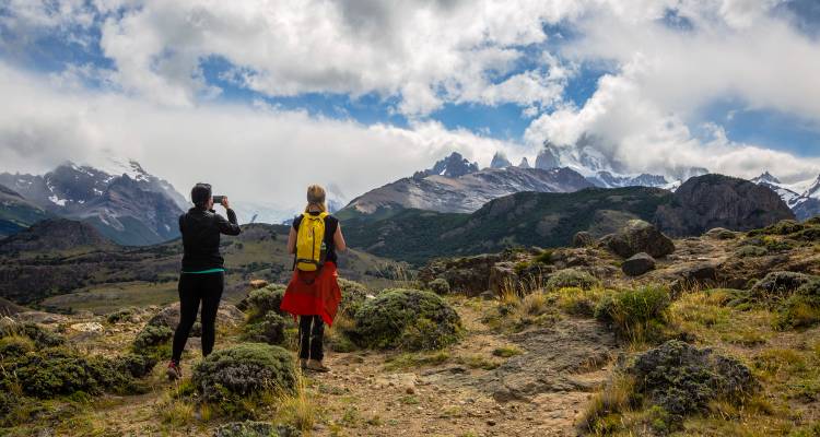 Zwei Wanderer halten auf einem felsigen Pfad an, um das zerklüftete, in Wolken gehüllte Mount Fitz Roy-Massiv zu fotografieren.