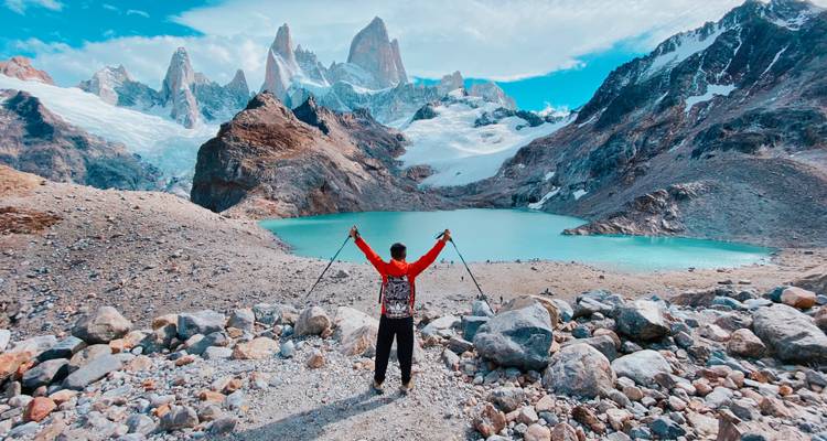 Triumphierender Wanderer hebt Stöcke neben einem türkisfarbenen Gletschersee unter dem dramatischen Fitz-Roy-Massiv.
