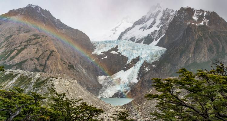 Un arc-en-ciel vibrant s'étend à côté d'un glacier suspendu qui se déverse dans un lac turquoise au milieu de pics rocheux escarpés.