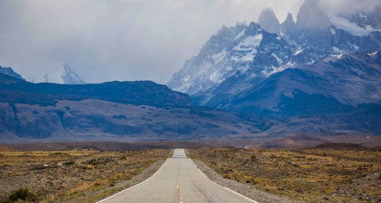 Eine gerade, leere Straße erstreckt sich in Richtung schneebestäubter Granitspitzen, die sich über die patagonische Steppe erheben.