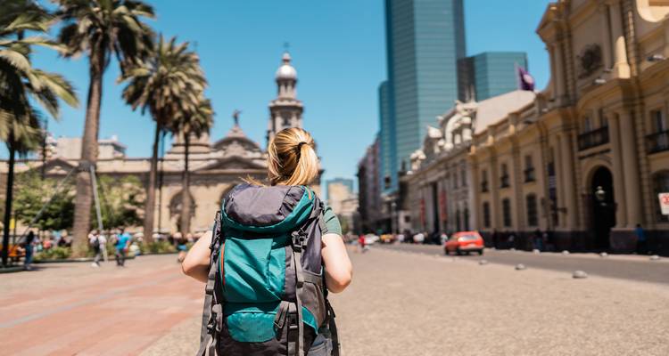 Rucksacktourist mit großem Rucksack läuft eine palmengesäumte Straße entlang, vorbei an historischen und modernen Gebäuden in Santiago.