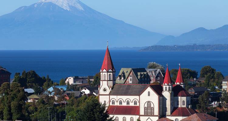 Escena pintoresca a orillas del lago con iglesia de torres rojas y majestuoso volcán nevado de fondo.