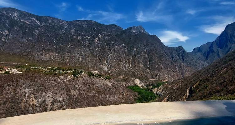 Amplio valle montañoso con pendientes áridas empinadas capturado desde un punto elevado sobre el borde de una piscina infinita bajo un cielo azul brillante.