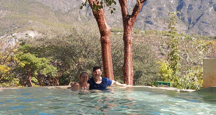 Pareja sonriente relajándose en una piscina infinita natural enmarcada por árboles y montañas escarpadas en el fondo.