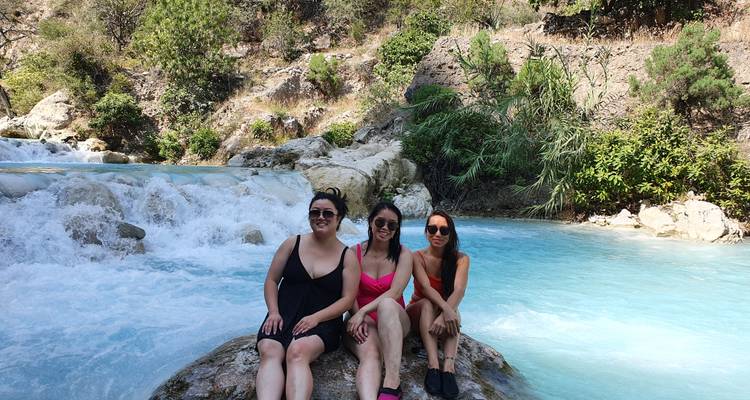 Tres amigos en trajes de baño sentados en una roca junto a un río turquesa que fluye rápidamente en un entorno de cañón exuberante.
