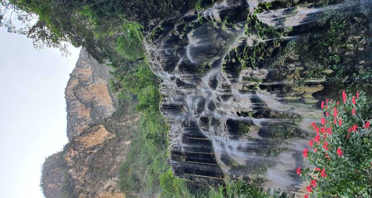 Cascada de varios niveles que desciende por paredes rocosas cubiertas de musgo verde, rodeada de follaje tropical y acantilados distantes.