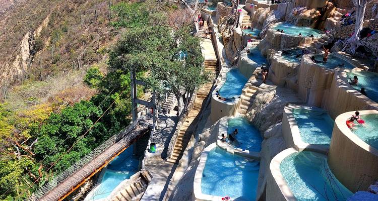 Vista de dron de terrazas en acantilado llenas de piscinas minerales de color azul vívido conectadas por escaleras y senderos entre árboles.