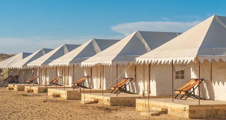 Row of luxurious canvas tents with wooden deck chairs set on golden desert sand under a blue sky.