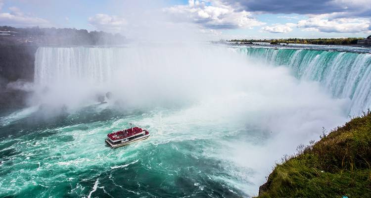 Krachtige Niagara Falls met nevel die hoog opstijgt terwijl een rondvaartboot vol roodgeklede passagiers de Horseshoe Falls nadert.