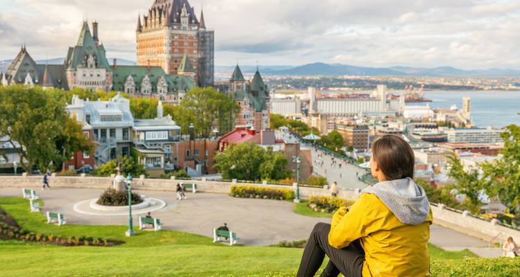 Reiziger in een gele jas kijkt uit over Château Frontenac in Quebec City en de Saint Lawrence rivier vanuit een grasrijk park.