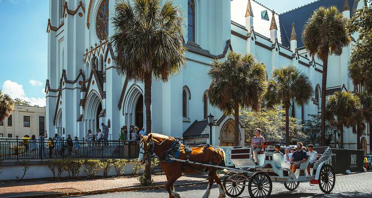 Un carruaje tirado por caballos con viajeros pasa por una iglesia blanca entre palmeras en Charleston.
