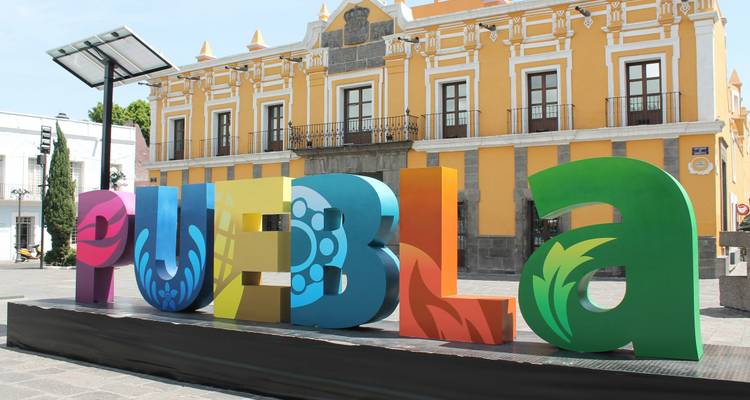 Letrero colorido en 3D de PUEBLA frente a un edificio colonial amarillo bajo un cielo soleado.