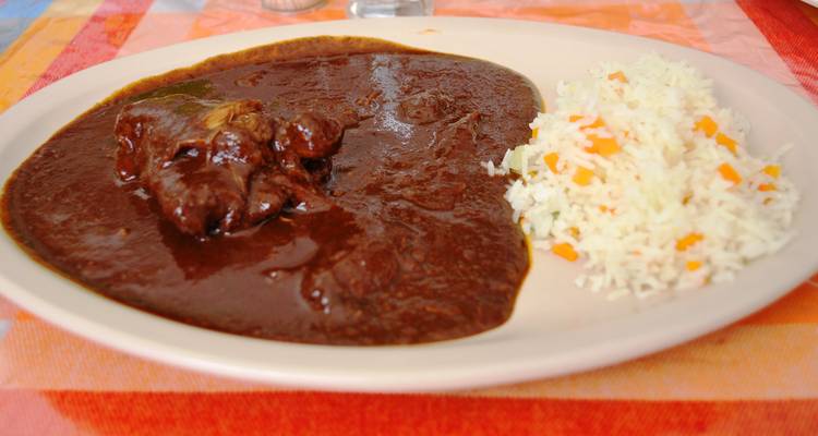 Un plato de rica salsa de mole oscuro con trozos de carne tierna y una porción de arroz con zanahorias cortadas en cubitos en una bandeja beige.