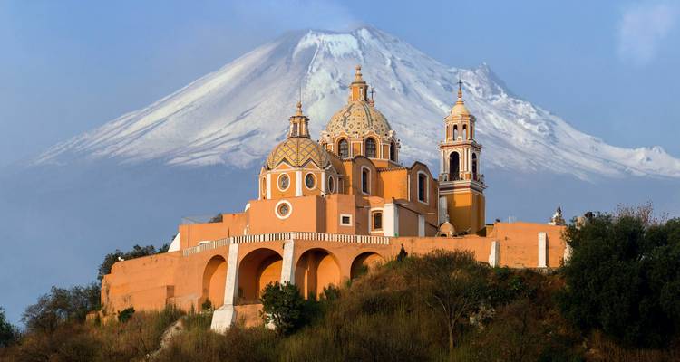 Una iglesia de color ocre en la cima de una colina se alza ante el volcán Popocatépetl cubierto de nieve bajo un cielo azul despejado.