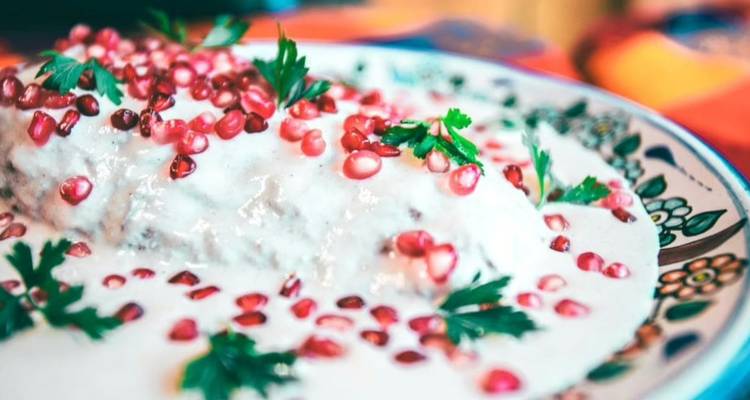 Primer plano de un chile en nogada tradicional cubierto con salsa cremosa de nuez salpicada con semillas de granada en un plato decorativo.