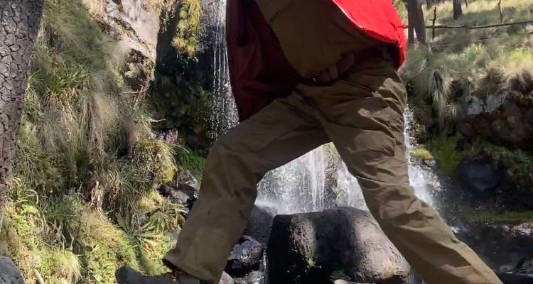 Ein Wanderer in einer roten Jacke schreitet über einen felsigen Bach neben einem kleinen Waldwasserfall.