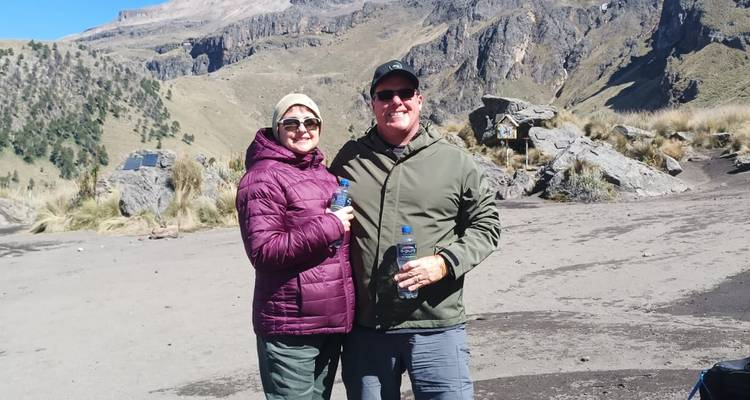 Ein lächelndes Paar in warmer Outdoor-Ausrüstung steht auf einer vulkanischen Ebene mit schroffen Gipfeln hinter ihnen.