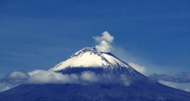 Der schneebedeckte Vulkan Popocatépetl stößt eine weiße Rauchfahne über Wolkenbändern vor einem tiefblauen Himmel aus.