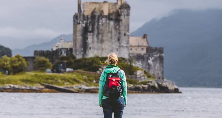 Een wandelaar met groene jas en rode rugzak kijkt over het water naar Eilean Donan Castle te midden van mistige bergen.