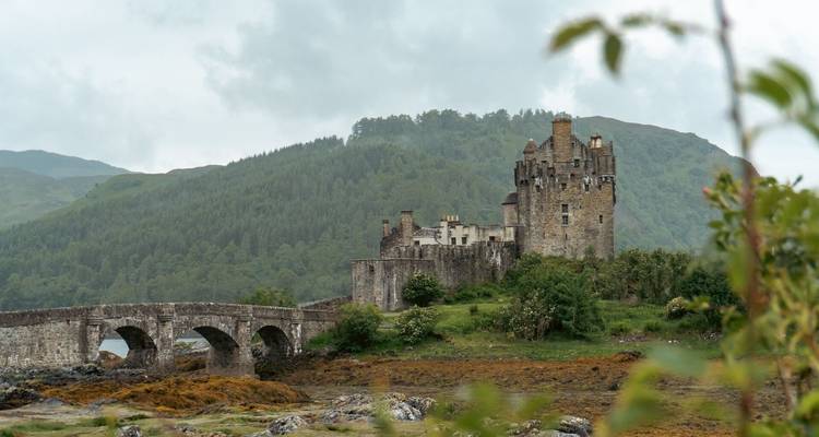 Stenen brug en Eilean Donan Castle liggen tegen beboste heuvels onder een bewolkte hemel.
