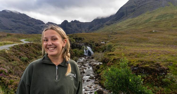 Een glimlachende vrouw poseert met ruige Schotse bergen en een kleine waterval achter haar.
