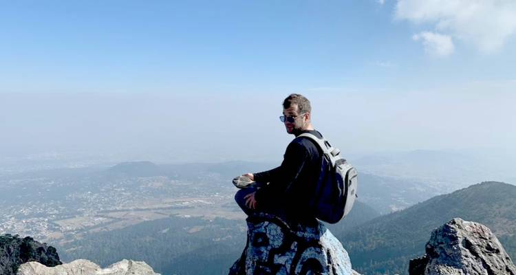 Junger Wanderer mit Rucksack sitzt auf einem felsigen Gipfel mit Blick auf dunstige Täler und entfernte Berge unter einem klaren blauen Himmel in der Nähe von Mexiko-Stadt.