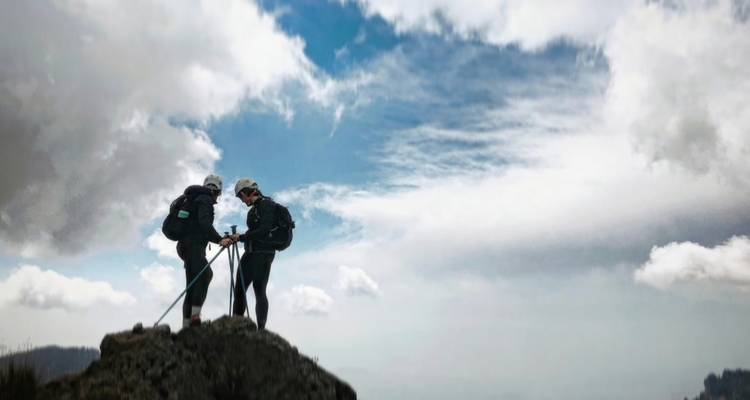 Zwei Wanderer mit Stöcken stehen auf einem felsigen Gipfel, der sich gegen dramatische Wolken und blauen Himmel abzeichnet.