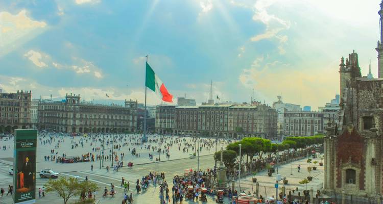 Weitläufige Ansicht des Zócalo-Platzes von Mexiko-Stadt mit riesiger mexikanischer Flagge, historischen Gebäuden und Menschenmengen unter dramatischen Himmelsstrahlen.