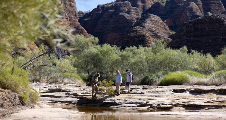 Drie wandelaars verkennen een rotsachtig beekbed onder de bijenkorven-vormige koepels van het Bungle Bungle Range.
