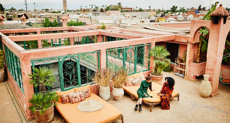 Twee vrienden kletsen op de zachte dakterrassen van een roze riad met uitzicht over de skyline van Marrakesh.