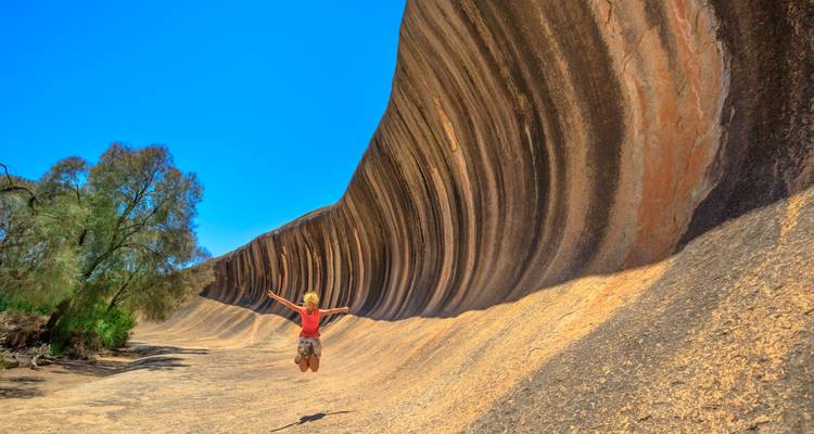 Kind springt fröhlich vor der aufragenden geschwungenen Granitwand des Wave Rock unter lebendigem blauen Himmel.