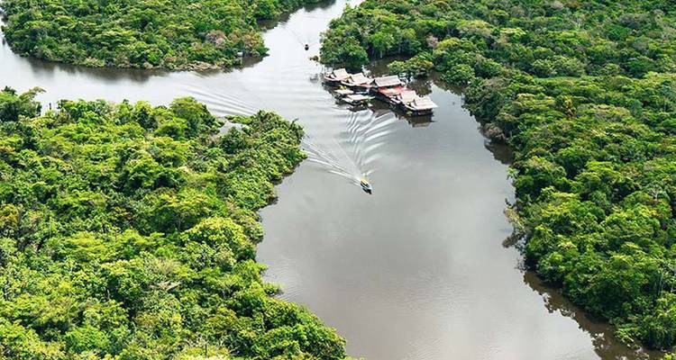 Luchtfoto van een kronkelende junglerivier met weelderig groen regenwoud en een speedboot die een wit kielzog trekt richting een drijvende lodge.
