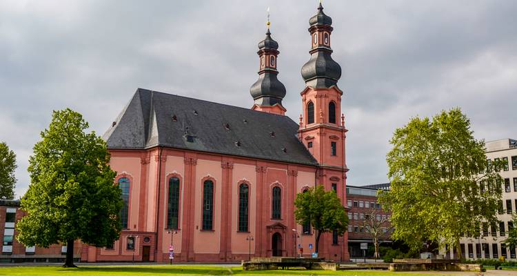 Eine rosafarbene Barockkirche steht vor einem grauen Himmel im deutschen Stadtzentrum.